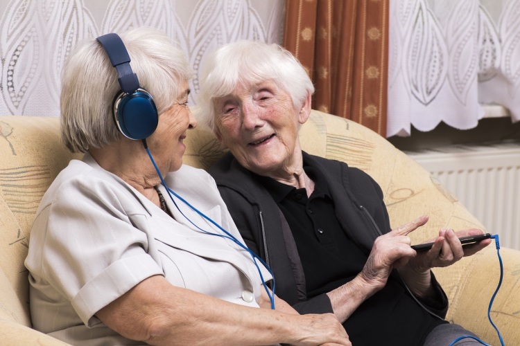 Two women, sitting on a sofa. One is wearing headphones, the other is holding a mp3 player. Both are smiling.